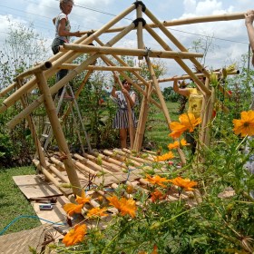a female standing on a ladder atop a bamboo framed building (under construction) in a pretty natural landscape setting with orange flowers in the foreground